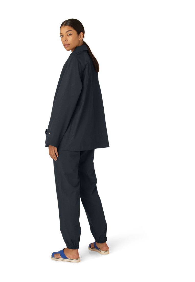 Giacca oversize di un blu scuro con colletto, abbinata a pantaloni slim coordinati. Il modello indossa sandali blu con suola testurizzata.