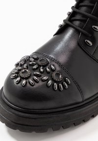 Czarna skórzana boot z teksturowanym noskiem ozdobionym metalowymi ćwiekami i dekoracyjnymi kamieniami. Posiada grubą podeszwę z głębokimi wzorami bieżnika.