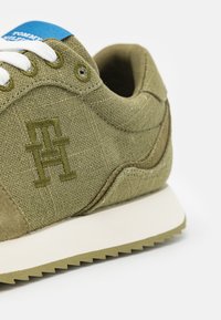 Sněrovací tenisky olivové barvy s bílými tkaničkami, vyšitým logem, modrým jazykem se štítkem, bílou podrážkou a vroubkovanou olivovou podešví.