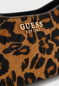 Dettaglio in tessuto con stampa leopardata e logo dorato "GUESS EST 1981" sul bordo di una scarpa o accessorio con finitura nera.