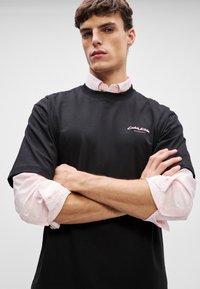 T-shirt preta de manga curta, feita de algodão, com um pequeno logótipo branco. Uma camisa rosa de manga longa é visível por baixo, com as mangas enroladas.