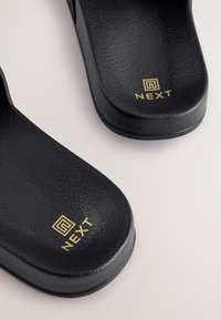Sandali con fascia neri in materiale testurizzato con il marchio "NEXT" dorato sulla superficie superiore. Bordi lisci e arrotondati.