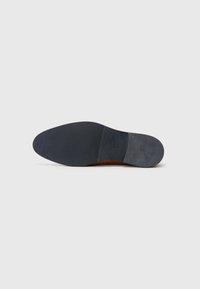 Suela de zapato de cuero con una sección superior marrón y una parte inferior de goma negra suave. Presenta una puntera redondeada y detalles de hardware mínimos.