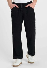 Man in zwarte corduroy broek met elastische tailleband en trekkoord, handen in zijn zakken, gecombineerd met witte sneakers met rubberen zolen.