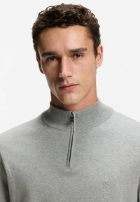 Szary dzianinowy sweter z wysokim kołnierzem i półzamkiem. Posiada subtelne logo "BOSS" na piersi oraz ściągacze na mankietach. Miękka faktura.