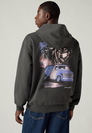 Homme portant un sweat à capuche sombre avec un graphique d'une camionnette bleue, un loup, un éclair et une lune dans le dos, debout devant un fond uni.