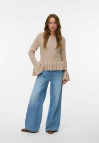 Maglione beige a coste con maniche a campana e orlo plissettato, abbinato a jeans a gamba larga azzurri e scarpe marroni. Sfondo semplice.