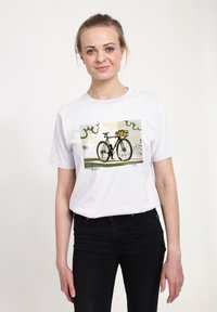 Žena v bílé tričku s grafikou vintage jízdního kola s košíkem, v černých džínách, stojící na neutrálním pozadí.