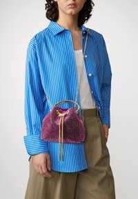 Violet sequined bucket bag dengan tali rantai emas dan penutupan tali serut, dipakai dengan kemeja bergaris biru yang memiliki potongan santai.
