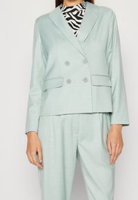 Vaaleansininen, kaksirivinen bleiseri, jossa on kaksi nappia ja sivutaskut, yhdistetty kuvioituun toppiin ja vastaaviin housuihin. Sileä kangaspinta.