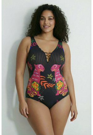 Frau mit lockigem Haar, die einen schwarzen Einteiler mit pinkfarbenen Tigern und bunten Blumenmustern trägt, lächelt vor einem schlichten Hintergrund.