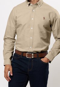 Beiges Hemd zum Knöpfen, aus Baumwollstoff, mit langen Ärmeln, weißen Knöpfen und einem kleinen Logo. Kombiniert mit dunkelblauen Jeans und einem braunen Gürtel.