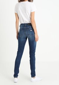 Vaqueros de mezclilla oscuros y ajustados con cintura media, bolsillos traseros y un pequeño logotipo. Combinados con una camiseta blanca de manga corta y zapatillas blancas.