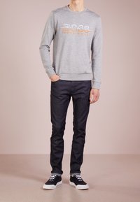 Šedá mikina s kulatým výstřihem a logem 'BOSS' v bílé a oranžové barvě, nošená s tmavými džíny a černými teniskami s bílou podrážkou.