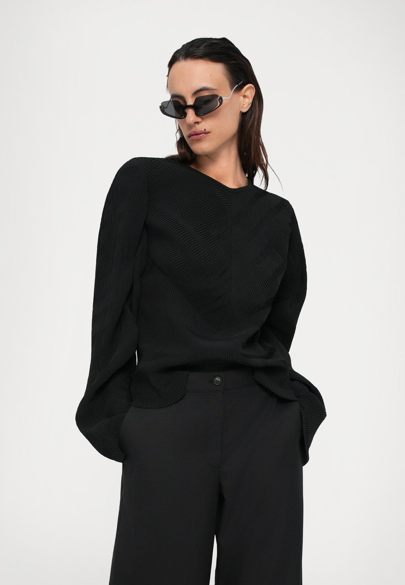 Femme portant un haut noir à manches longues texturé, un pantalon noir et de petites lunettes de soleil noires, les mains dans les poches, contre un fond clair uni.