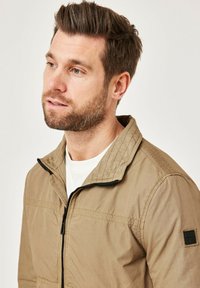 Khaki zip-up jacket with a stand collar, featuring textured fabric, black zipper, and a patch on the left sleeve. Smooth, lightweight design.