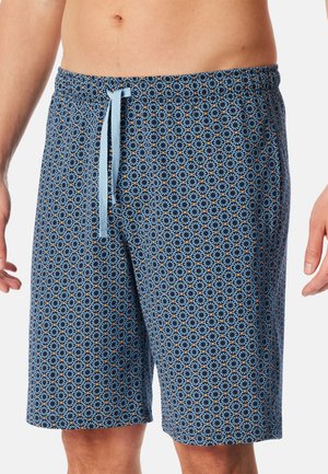 Mężczyzna w niebieskich szortach z wzorem geometrycznych kół z pomarańczowymi akcentami, z ściągaczem, przyciętych na wysokości talii i kolan.