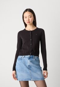 Črni suknjič z okroglim izrezom, dolgimi rokavi in sprednjim gumbom. Kombiniran z svetlo modrim denim krilom, ki prikazuje klasičen kroj in obliko.