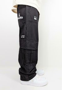 Schwarze Cargohosen mit mehreren Taschen, versehen mit weißen, gestickten Logos und Schriftzügen. Hergestellt aus strapazierfähigem Stoff mit lockerer Passform. Weiße Sneaker darunter.