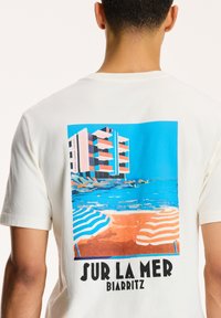 Balta medvilninė marškinėliai su spalvinga paplūdimio scena, kurioje pavaizduotas pastatas ir dryžuoti paplūdimio skėčiai. Tekstas sako "SUR LA MER BIARRITZ."
