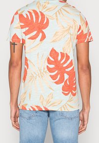 Kurzarmhemd mit hellblauem Hintergrund. Verfügt über große rot-beige tropische Blattmuster. Hergestellt aus leichtem Stoff.