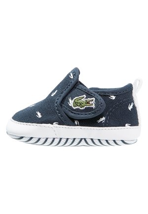 Blauwe canvas babyschoen met witte details, elastische bandsluiting en geborduurd krokodillenlogo. Rubberen zool met een gestreept patroon en textuur.