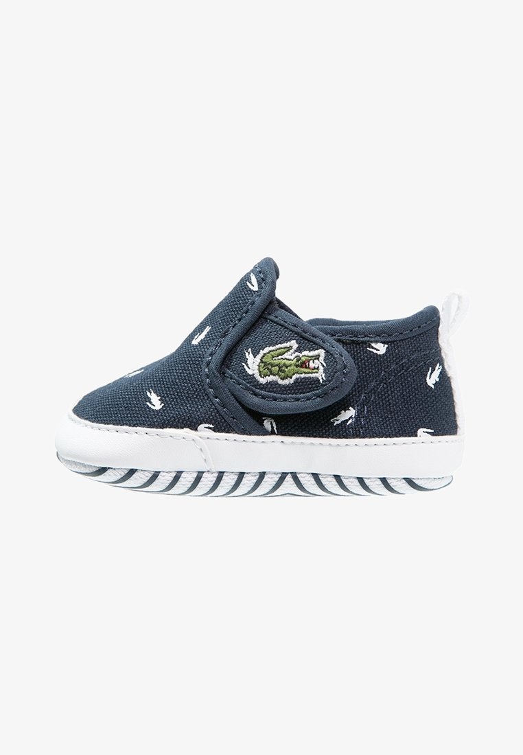 Chaussure bébé en toile bleue avec détails blancs, fermeture par sangle élastique et logo crocodile brodé. Semelle en caoutchouc texturée avec motif rayé.