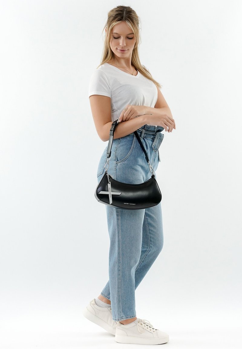 Borsa a tracolla in pelle nera con forma curvata, accentata da una croce argentata e tracolla regolabile. Indossata con jeans azzurri chiari e sneakers bianche.