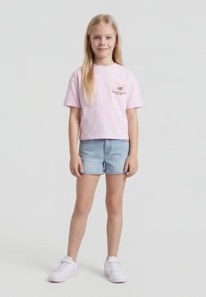 Νεαρή κοπέλα στέκεται χαμογελώντας, φορόντας ένα ανοιχτό ροζ T-shirt με λογότυπο, ανοιχτόχρωμα τζιν σορτς και λευκά αθλητικά παπούτσια, μπροστά σε απλό φόντο.