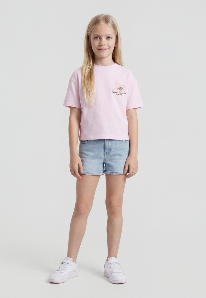 Νεαρή κοπέλα στέκεται χαμογελώντας, φορόντας ένα ανοιχτό ροζ T-shirt με λογότυπο, ανοιχτόχρωμα τζιν σορτς και λευκά αθλητικά παπούτσια, μπροστά σε απλό φόντο.