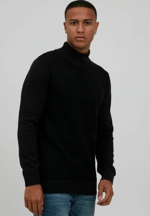 Schwarzer Reißverschluss-Pullover mit gerippter Textur, hohem Kragen und langen Ärmeln. Kombiniert mit blauen Jeans, die einen enganliegenden Schnitt haben.