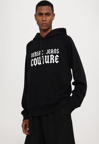 Μαύρη φούτερ μπλούζα φτιαγμένη από μαλακό ύφασμα, με λευκό κείμενο "VERSACE JEANS COUTURE". Ριπ περιλαίμια και κάτω άκρο, χαλαρή εφαρμογή με κουκούλα που έχει κορδόνι.