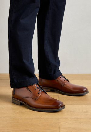 Chaussures habillées en cuir marron avec une finition lisse, design à lacets et petit talon empilé. Portées avec un pantalon bleu marine, mettant en valeur des ourlets roulés.