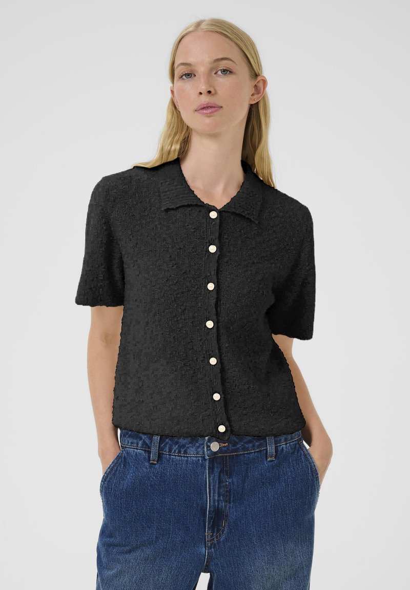 Femme aux longs cheveux blonds portant une chemise noire à manches courtes avec texture et un jean bleu, se tenant les mains dans les poches contre un fond blanc.