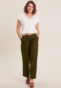 Weißes Kurzarmoberteil mit dekorativem Rundhalsausschnitt, kombiniert mit olivgrünen, hochgeschnittenen Hosen und braunen Plateausandalen.