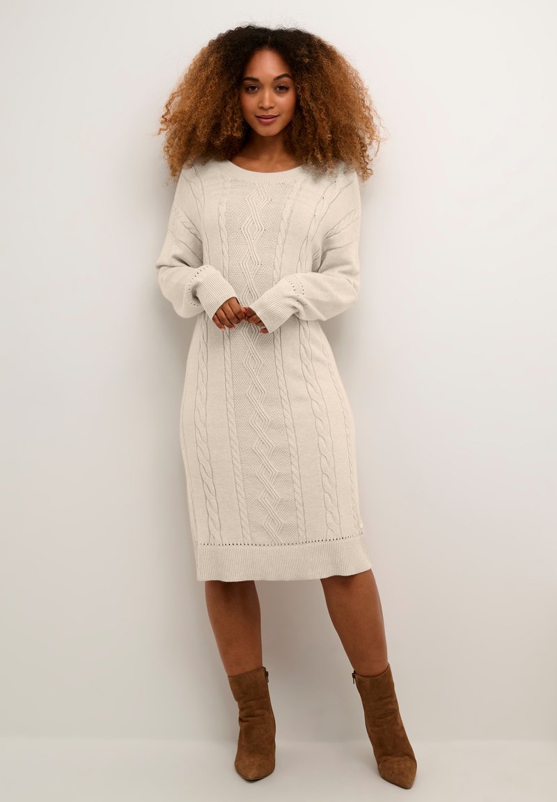 Rochie tricotată de culoare bej deschis, cu modele din tricot cu cablu, mânici și tiv cu linii ondulate. Design de lungime medie cu o croială lejeră.