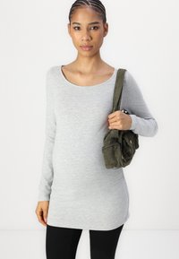Graues Langarmshirt mit rundem Ausschnitt, kombiniert mit schwarzen Leggings. Die Person hält eine strukturiertes grünes Cordura-Tasche.
