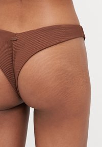 Brown ribbed bikini bottom featuring a smooth texture and a narrow waistband. Notable accent includes a small brand tag under the waistband.