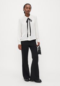 Blouse blanche à manches longues avec un nœud en ruban noir au col, associée à un pantalon noir taille haute et des chaussures à plateforme noires. Design simple.