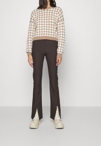 Stickad tröja med houndstooth-mönster i beige och vitt, tillsammans med mörkbruna utsvängda byxor med slitsar vid anklarna och kraftiga beiga sneakers.