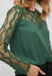 Top verde con tessuto a tinta unita e maniche in pizzo verde intricati. Le maniche presentano un motivo floreale e una texture semi-trasparente.