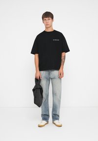 Zwart T-shirt met korte mouwen en witte tekst, gecombineerd met lichtblauwe spijkerbroek en crèmekleurige sneakers. Een zwarte jas wordt in de linkerhand vastgehouden.