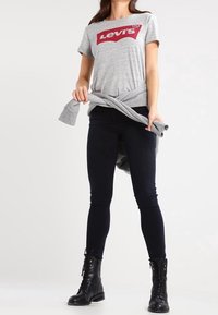 Camiseta gris de manga corta con el logo rojo de Levi's, anudada en la cintura, combinada con jeans ajustados oscuros y botines negros con cordones.