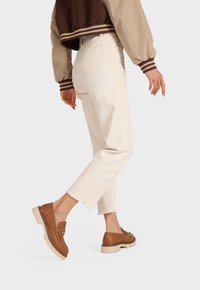 Mocassini in pelle marrone con dettaglio in catena dorata, jeans aderenti crema con orli sfilacciati e una giacca bomber marrone con strisce.