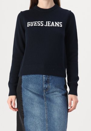 Kobieta ubrana w granatowy sweter z białym napisem "GUESS JEANS" oraz dwukolorową spódnicę dżinsową z czarnymi panelami po bokach.