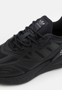 Svart atletisk sko med en mesh-ovandel, texturerade överlager, tjocka snören och en mönstrad sula med Adidas-logotypen på tungan.