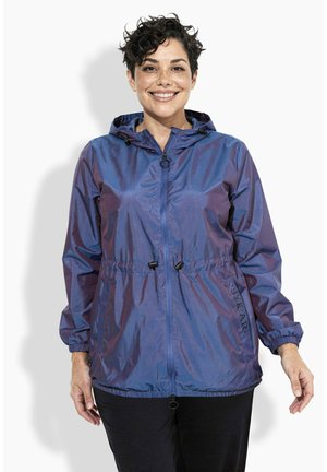 Lächende Person mit kurzen lockigen Haaren, die eine glänzende lila Kapuzenjacke mit Frontreißverschluss sowie verstellbarem Bund und Bündchen trägt.