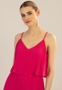 Plissiertes pinkes Kleid mit V-Ausschnitt, geschichteter Gestaltung und dünnen Trägern mit goldenen Akzenten. Sanfte Textur mit fließender Silhouette.