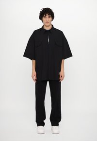 Black short-sleeve shirt with two chest pockets, button closure, and loose fit, paired with black pants featuring white stitching and white sneakers.