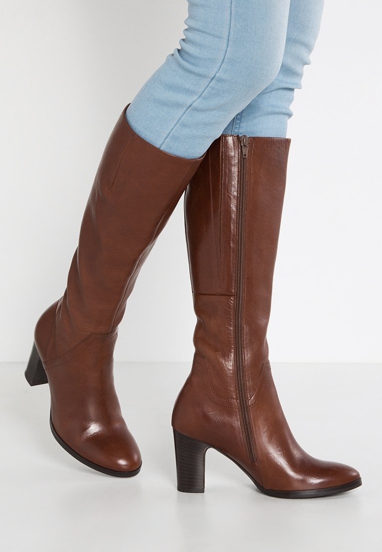 Bottes hautes jusqu'au genou en cuir marron avec une finition lisse, une fermeture éclair latérale et un talon épais. Associées à un jean ajusté bleu clair.